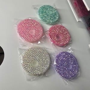May include: Five round, bedazzled cup lids in various colors: light pink, hot pink, mint green, white, and purple. Each lid is covered in small, sparkling rhinestones and packaged in a clear plastic bag.