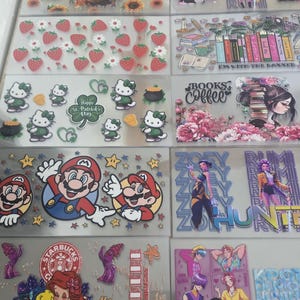 May include: A collection of clear stickers featuring various designs. Designs include a Highland cow with sunflowers, strawberries, Hello Kitty, Mario characters, and characters from Disney's The Little Mermaid. Other stickers feature text and illustrations.