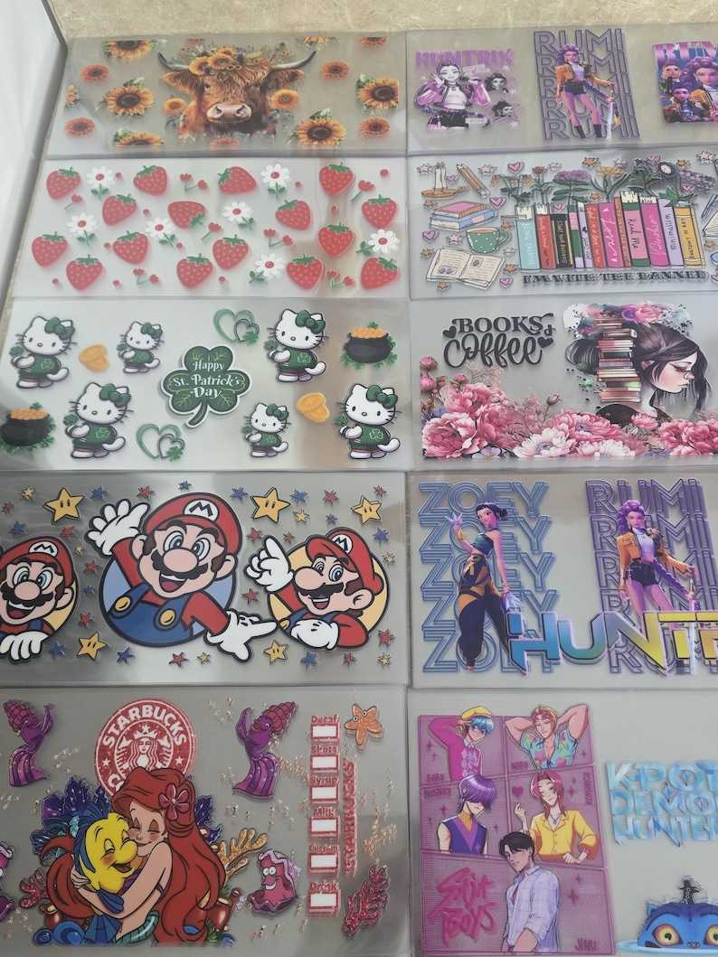 May include: A collection of clear stickers featuring various designs. Designs include a Highland cow with sunflowers, strawberries, Hello Kitty, Mario characters, and characters from Disney's The Little Mermaid. Other stickers feature text and illustrations.