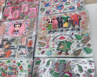 May include: A collection of clear plastic pouches filled with colorful stickers. The stickers feature various designs, including cartoon characters, holiday themes, and inspirational quotes. Some pouches have text like "FRIENDS" and "YOU CAN DO IT!"