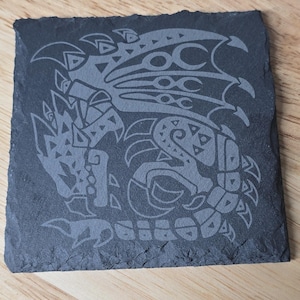 May include: Square, dark gray slate coaster with a detailed, etched design of a stylized dragon or monster. The design features intricate patterns and shapes, likely for a video game or fantasy theme. The coaster sits on a wooden surface.