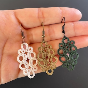 Puede incluir: Tres pares de delicados pendientes de ganchillo en blanco, dorado y verde. Cada pendiente presenta un diseño único e intrincado con múltiples bucles y círculos.