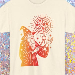 May include: Cream-colored t-shirt featuring a wizard with a long beard and a pointed hat, holding a wand. The wizard is conjuring a pizza with mushrooms and pepperoni. The design is in shades of orange and red.