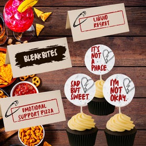 Op de afbeelding: Een houten tafel met eten en drinken, waaronder cupcakes met toppers. De toppers lezen "It's Not A Phase", "Sad But Sweet" en "I'm Not Okay". Andere borden zeggen "Bleak Bites", "Emotional Support Pizza" en "Liquid Regret".