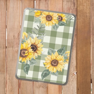 Puede incluir: Una funda transparente para tableta con un diseño de acuarela de girasoles amarillos y hojas verdes sobre un patrón de cuadros vichy verde claro y blanco. La funda es rectangular y tiene un borde transparente.
