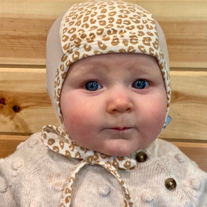 May include: A baby wearing a beige knitted sweater with brown buttons and a beige and brown leopard print bonnet with a matching tie.
