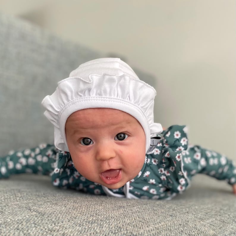 May include: A baby wearing a white ruffled bonnet and a green and white floral onesie. The baby is lying on its stomach and looking directly at the camera.