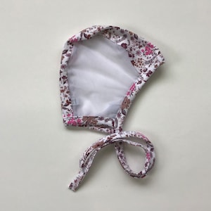 May include: A white baby bonnet with a floral pattern in shades of pink, brown, and green. The bonnet has a white mesh lining and ties with fabric strings. The floral design covers the exterior of the bonnet.