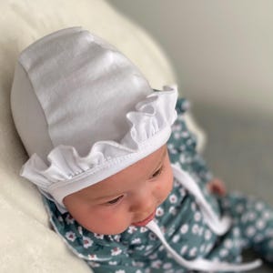 May include: A white baby bonnet with a ruffled edge. The bonnet is made of soft cotton and is perfect for keeping a baby's head warm.