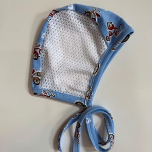 May include: A baby bonnet with a light blue fabric featuring a pattern of red and brown bicycles. The bonnet has a white mesh interior and light blue ties for securing. The edges are trimmed with light blue fabric.
