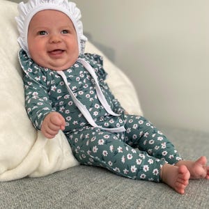 May include: A baby wearing a white bonnet and a green floral jumpsuit with white flowers. The baby is sitting on a green and white patterned blanket.