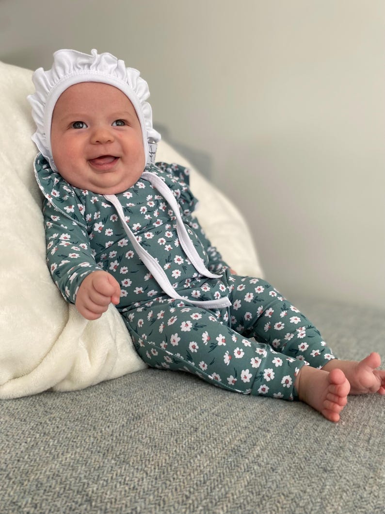 May include: A baby wearing a white bonnet and a green floral jumpsuit with white flowers. The baby is sitting on a green and white patterned blanket.