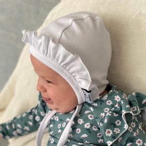 May include: A white baby bonnet with a ruffled edge. The bonnet has a small label that says "Emile et Rose".