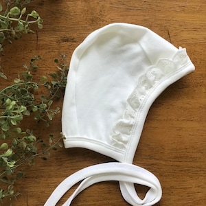 May include: White baby bonnet with a lace trim and ties. The bonnet is made of soft cotton and is perfect for a newborn baby.