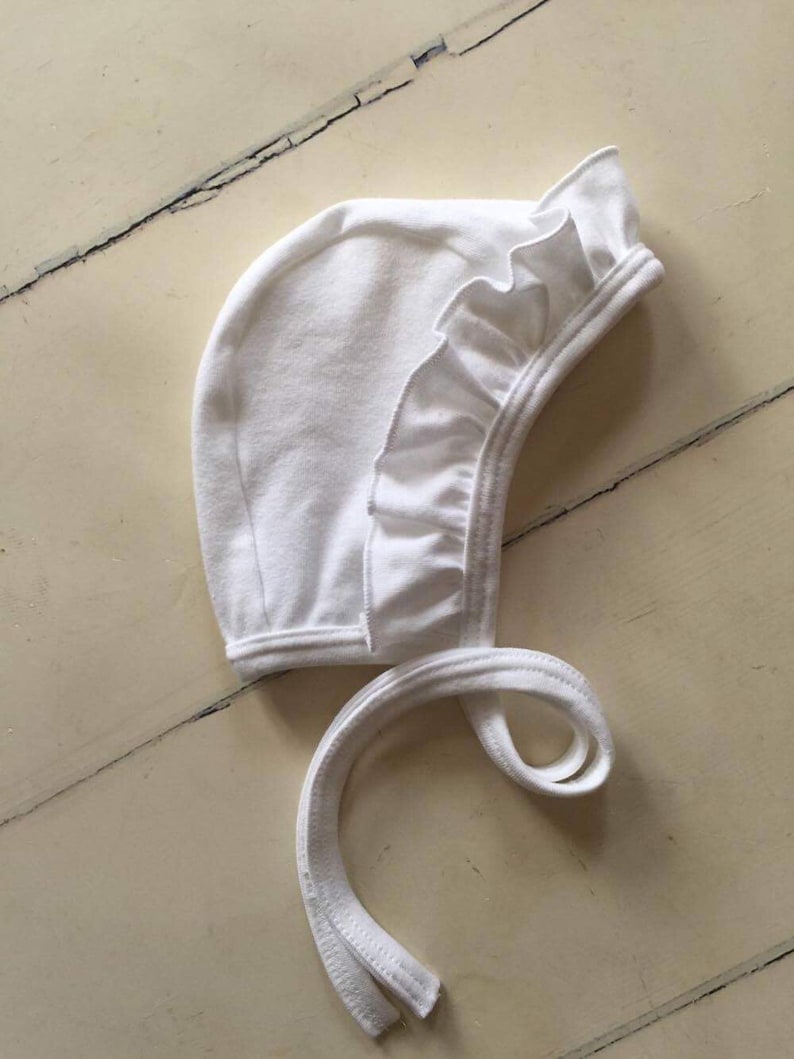 May include: White baby bonnet with a ruffled edge and ties. The bonnet is made of soft cotton and is perfect for keeping a baby's head warm and protected.