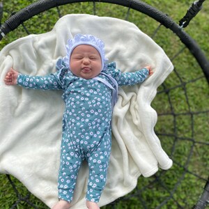 May include: A baby wearing a white bonnet and a teal floral jumpsuit is lying on a white blanket in a black swing.