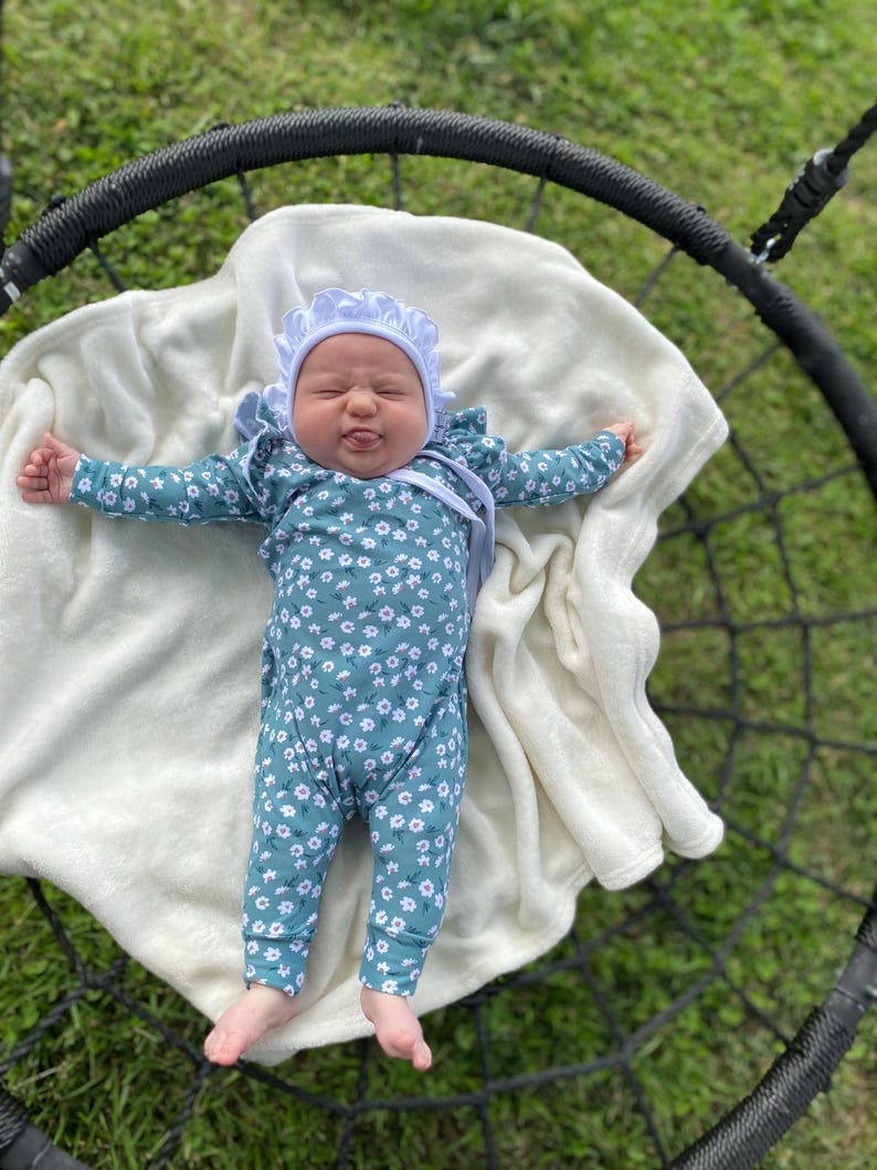 May include: A baby wearing a white bonnet and a teal floral jumpsuit is lying on a white blanket in a black swing.