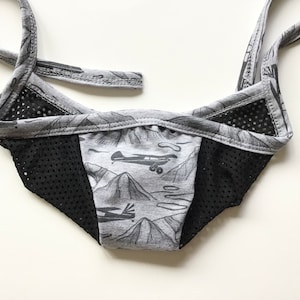 Könnte beinhalten: Graue und schwarze Stoffmaske mit einem Mesh-Panel und einem Aufdruck von Flugzeugen und Bergen.