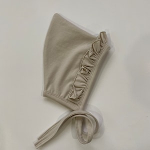 May include: A beige baby bonnet with a ruffled edge and a long tie. The bonnet is made of a soft, stretchy fabric.