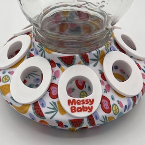 Peut inclure: Un bocal en verre transparent est posé sur une base blanche avec six ouvertures ovales. La base est décorée d'un motif de fruits colorés, dont des pastèques, des fraises et des citrons. Le texte "Messy Baby" est imprimé en rouge sur la base.