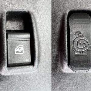 May include: Two black car interior buttons. The left button has a window and arrow symbol. The right button has a stylized bird design. Both buttons are set in a dark gray panel.