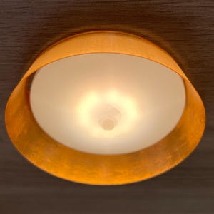 Könnte beinhalten: Eine kreisförmige Deckenleuchte mit einem warmen, metallisch kupferfarbenen Außenrand und einer zentralen, mattweißen Glaskuppel, die ein sanftes, diffuses Licht abgibt und ein fokussiertes, kreisförmiges Beleuchtungsmuster erzeugt.