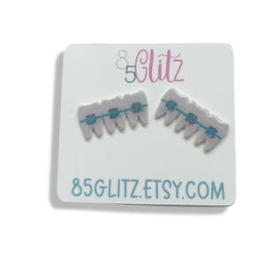 May include: A pair of novelty earrings shaped like teeth with braces, presented on a white card. The earrings are light purple with blue braces. The card has the text "85 Glitz" and "85GLITZ.ETSY.COM" in teal.