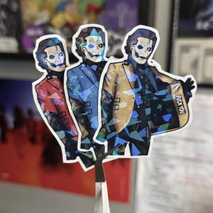 May include: A set of three die-cut stickers featuring a figure with white face paint and dark eye makeup, wearing a suit jacket in red, blue, or gold. The stickers have a holographic finish with geometric shards and the text FUCK YOU.