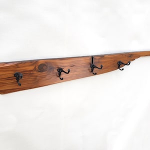 May include: A rustic wooden coat rack with four black metal hooks. The wood is a rich brown color with a natural, weathered finish. The rack is mounted on a white wall.