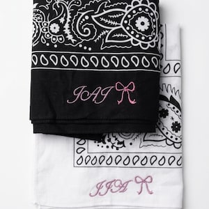 May include: Two folded bandanas, one black and one white, with paisley patterns. The black bandana has pink embroidered text that reads "JAI R". The white bandana has pink embroidered text that reads "JJA R".