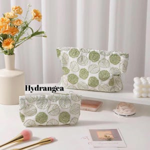 Puede incluir: Dos neceseres blancos con un estampado de hojas verdes. Las bolsas están bordadas con un diseño de hojas y la palabra "Hydrangea" está impresa en la imagen. También se ven brochas de maquillaje y un jarrón con flores.