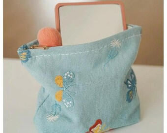 May include: A light blue fabric pouch with embroidered floral designs, holding a small mirror and a peach-colored makeup brush. The pouch has a drawstring closure and is made of a soft, textured material.
