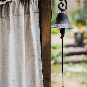 Può includere: Una campana da parete in metallo scuro con un motivo decorativo a forma di uccellino e foglie sulla parte superiore. La campana pende da una staffa curva fissata a un palo di legno scuro, con un lungo cordino nero e un battacchio sottostante.
