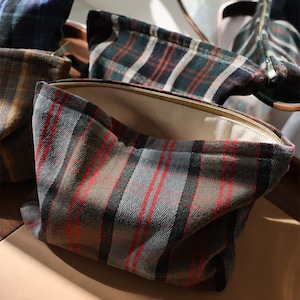 May include: A zipped pouch featuring a grey, black, and red plaid pattern with a herringbone texture. The interior is lined with a solid cream-coloured fabric. The pouch has a rectangular shape with a top zip closure and soft, flexible sides.