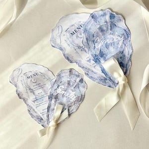 May include: Oyster-shaped wedding menu cards with watercolor blue and gray designs. Each card features a name in white script and is tied with a cream ribbon. The menu includes starter, main, and dessert options.