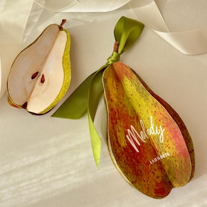 Peut inclure: Un objet décoratif en forme de poire avec une tige et un ruban verts, et une demi-poire coupée. L'objet en forme de poire porte le mot "Melody". Les couleurs sont le jaune, le rouge et le vert.