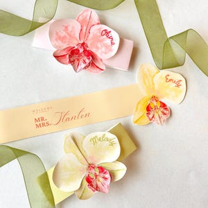 May include: Three orchid-shaped place cards with names written on them, along with a banner that reads "Mr. & Mrs. Stanton". The orchids are pink and yellow with red and white details. Green ribbon is also present.