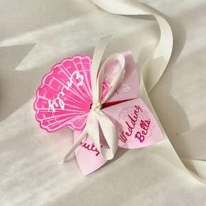 Op de afbeelding: Een roze huwelijksbedankje in de vorm van een bloem, met de naam "Emily" gedrukt op een waaiervormig bloemblad. Het bedankje is vastgebonden met een wit lint en bevat de woorden "Wedding Bells".