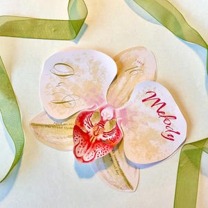 May include: An orchid-shaped wedding place card with the name "Melody" in red script. The card has a cream and pink color scheme, with the text "Table 16" and wedding details. A green ribbon is draped around the card.