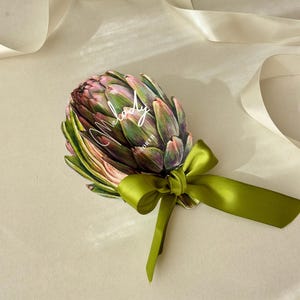 Puede incluir: Una tarjeta decorativa con forma de alcachofa con la palabra "Melody" en escritura blanca. La tarjeta presenta tonos verdes y rosas, con una cinta de raso verde atada en un lazo. Hay cintas en el fondo.