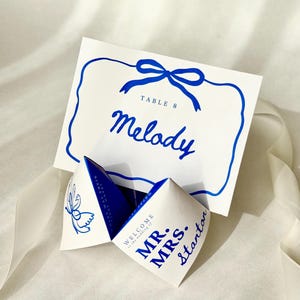 Puede incluir: Tarjetas de mesa de boda blancas con detalles azules. Una tarjeta presenta el nombre "Melody" y "Table 8" en escritura azul. También hay un adivino de papel plegado, con "Mr. Mrs. Stanton" impreso.