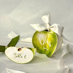 Può includere: Segnaposto a forma di mela verde con nastro bianco e testo. Una carta dice "Olivia" e l'altra "Tavolo 6". I segnaposto a forma di mela sono un design unico per matrimoni o eventi.