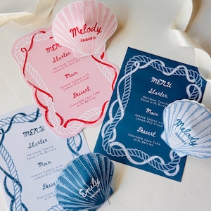 May include: A collection of custom menu cards and seashell-shaped place cards. The menus are in pink, white, and blue, with a nautical rope border. The place cards feature names like "Melody" and "Emily".