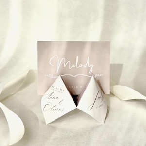 Peut inclure: Une décoration de table de mariage avec une carte rose blush portant l'inscription "Melody" en écriture élégante. Deux cartes blanches en forme de losange sont ouvertes, affichant les noms "Anna & Oliver" et la date "07.2026". Un ruban blanc est également présent.