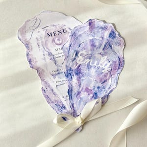 May include: Two oyster-shaped wedding menu cards with watercolor purple and blue designs. One card reads "MENU" and lists menu items. The other card has the name "Emily" in white script. A white ribbon is tied around the cards.