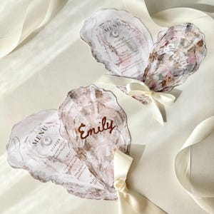 Può includere: Biglietti per il menu di nozze a forma di conchiglia di ostrica con un accento di perla e il nome "Emily" in oro. I biglietti sono legati con nastri color avorio e il menu include antipasti, portate principali e dessert. Il tema generale è elegante e nautico.