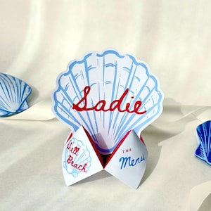 May include: A seashell-shaped place card with the name "Sadie" in red script. The card is blue and white with "Shell Beach" and "The Menu" printed on the folded sections. Additional seashell decorations are visible.
