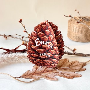 Könnte beinhalten: Ein dreidimensionales Tannenzapfen-Ornament aus Papier mit braunen Schuppen und dem weißen Schriftzug Enna und SWEENEY auf der Vorderseite. Das Ornament ist zusammen mit einem flachen Papier-Eichenblatt, kahlen Zweigen und braunem Garn abgebildet.