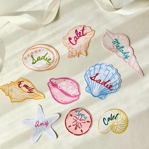 May include: A selection of seashell-shaped stickers, each bearing a name. The stickers are in shades of pink, orange, blue, and yellow. Names such as "Sadie", "Calet", "Melody", "Amy", "Tijra", and "Emily" are visible.