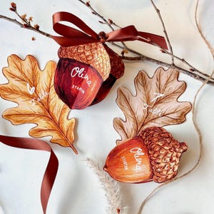May include: Two paper acorn-shaped place cards with brown ribbon, featuring the white printed text Olivia Stanton. One acorn is paired with a golden-brown oak leaf labeled Table 6, and the other with a light brown oak leaf labeled Table 7.